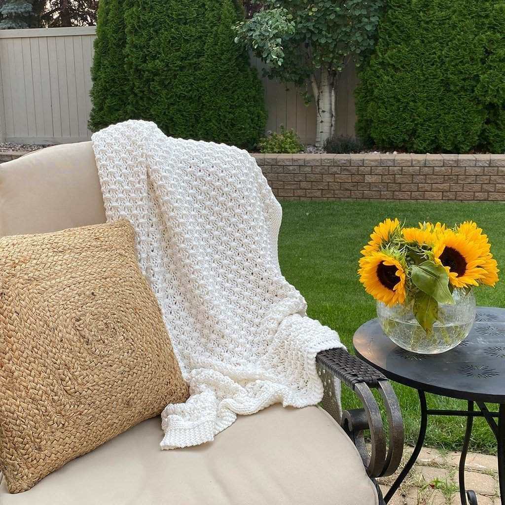 White crocheted blanket draped over a beige chair with a woven pillow, accompanied by a small table with sunflowers outdoors.