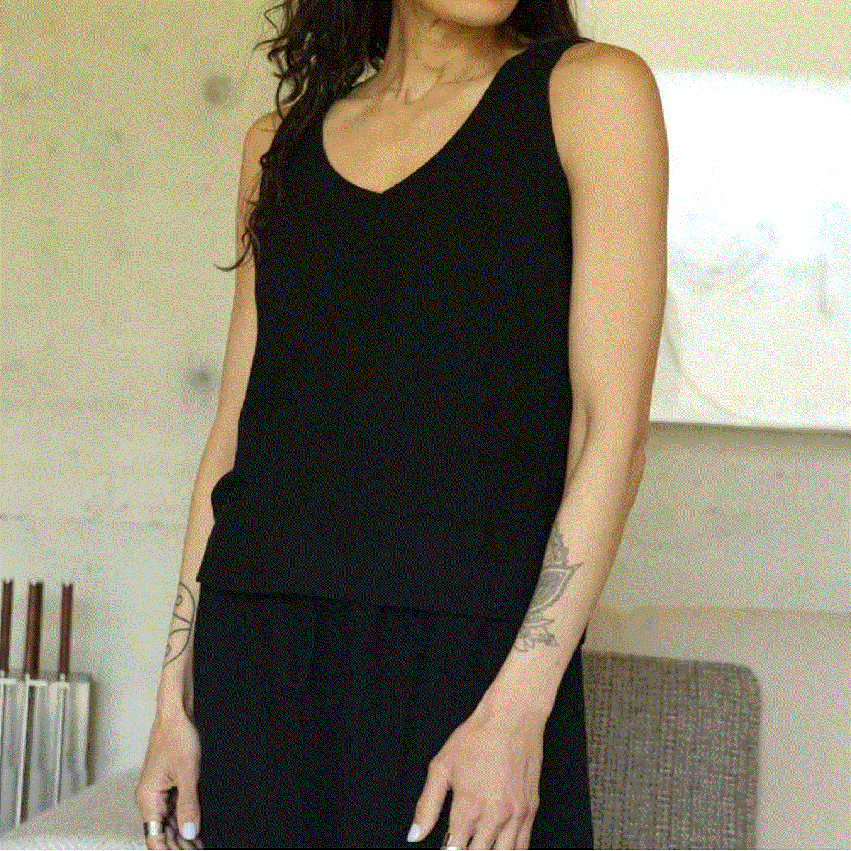 A woman wearing a black sleeveless V-neck top and black bottoms, standing indoors against a light neutral wall.