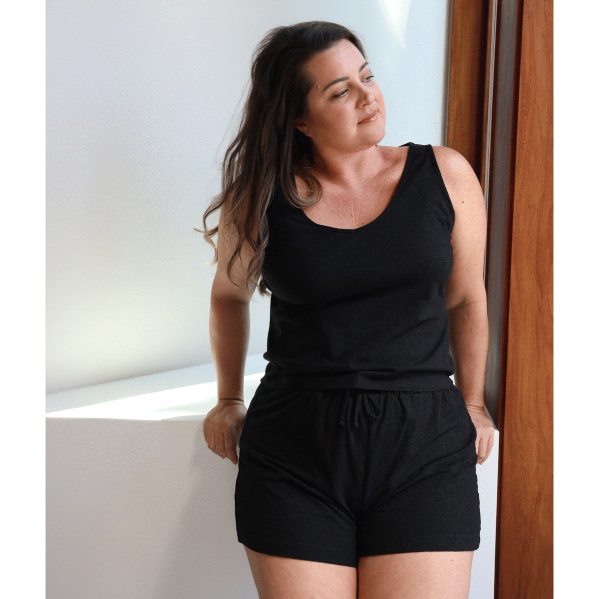 Woman wearing a black sleeveless top tank and shorts standing against a white wall.