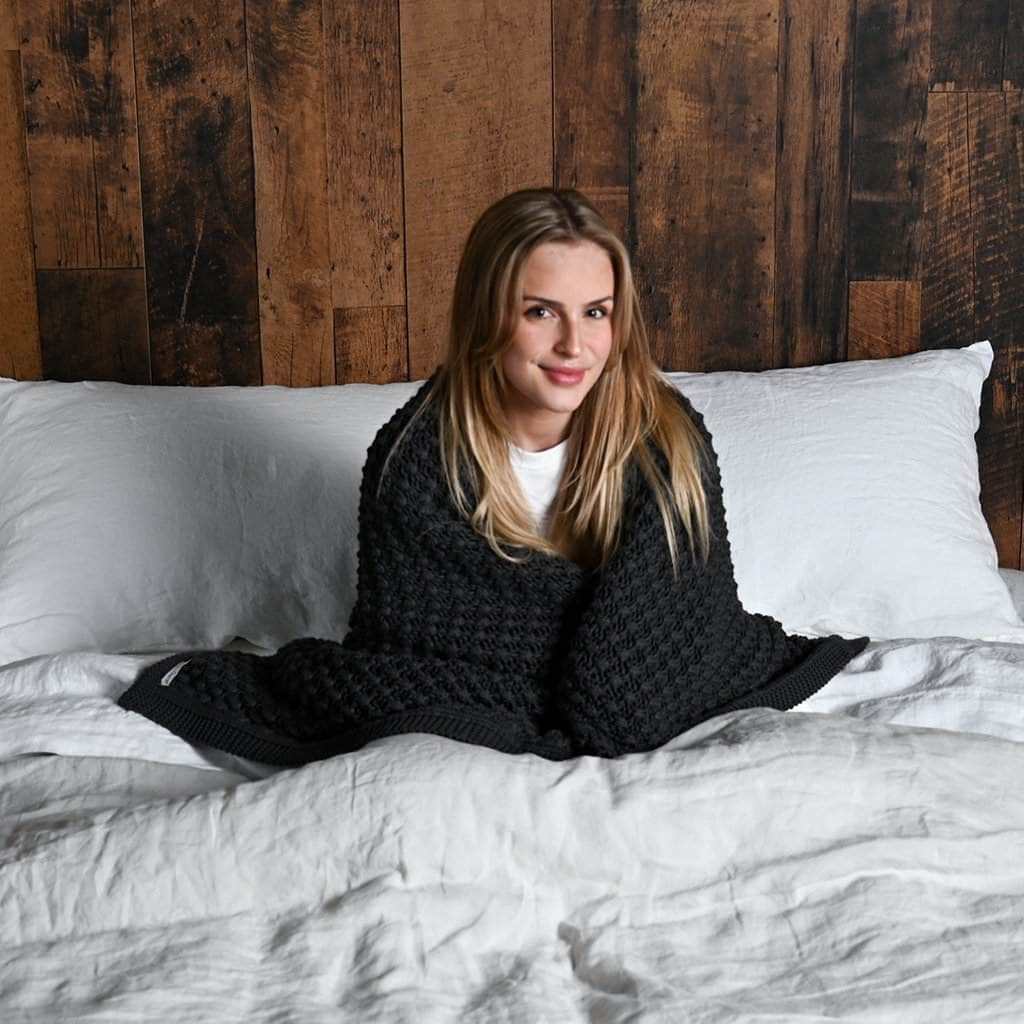 Woman sitting on a bed with a wooden paneled wall in the background caring knit blanket around shoulders 